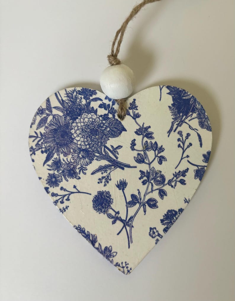 Op de afbeelding: Houten ornament in de vorm van een hart met een blauw bloemmotief op een cr&egrave;mekleurige achtergrond. Het ontwerp toont verschillende bloemen en bladeren. Een witte houten kraal is aan de bovenkant bevestigd met een natuurlijke touwlus om op te hangen.