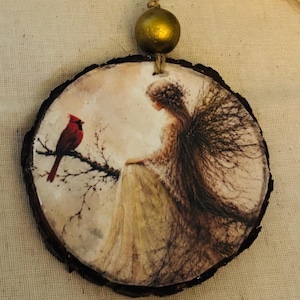 May include: A circular wood slice ornament featuring a printed illustration of a figure with dark, branch-like wings standing near a bright red cardinal perched on a thin twig. The ornament is suspended by a jute twine string with a gold bead.