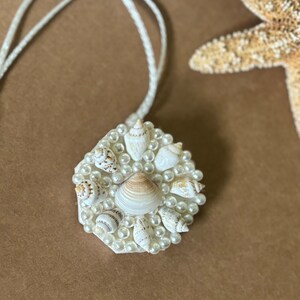 May include: A white hexagonal pendant necklace with a cluster of small white seashells and white pearls. The necklace is on a white cord.