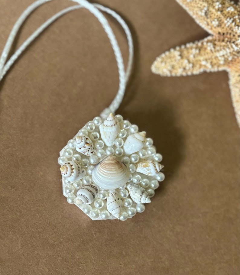 May include: A white hexagonal pendant necklace with a cluster of small white seashells and white pearls. The necklace is on a white cord.