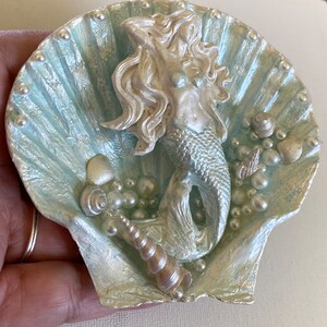 K&ouml;nnte beinhalten: Eine wei&szlig;e und blaue Muschel mit einer silbernen Meerjungfrauenfigur im Inneren. Die Muschel ist mit Perlen und Muscheln verziert.