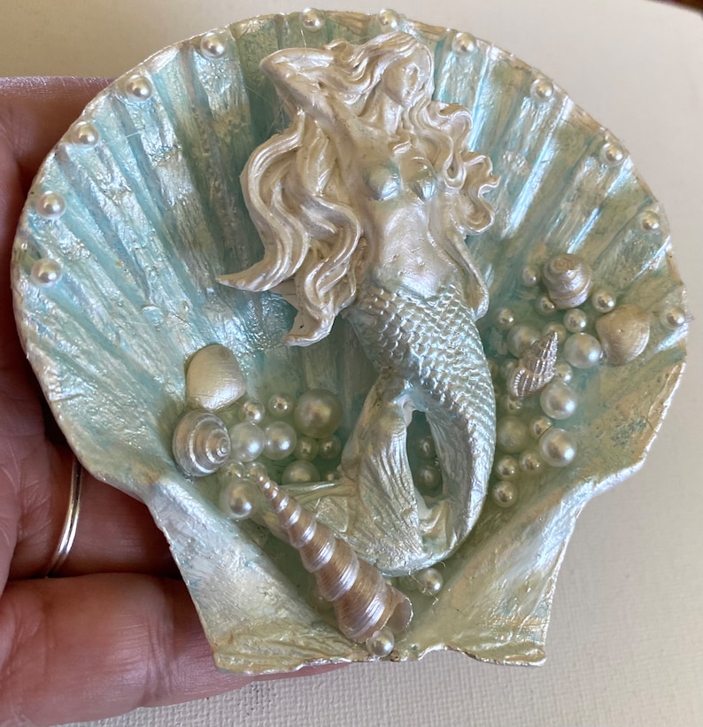 K&ouml;nnte beinhalten: Eine wei&szlig;e und blaue Muschel mit einer silbernen Meerjungfrauenfigur im Inneren. Die Muschel ist mit Perlen und Muscheln verziert.