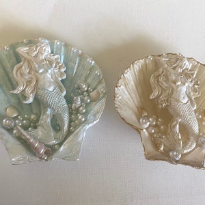 K&ouml;nnte beinhalten: Zwei dekorative Muschelschalen mit einer Meerjungfrauenfigur im Inneren. Eine Schale ist hellblau mit silbernen Akzenten und die andere ist wei&szlig; mit goldenen Akzenten.