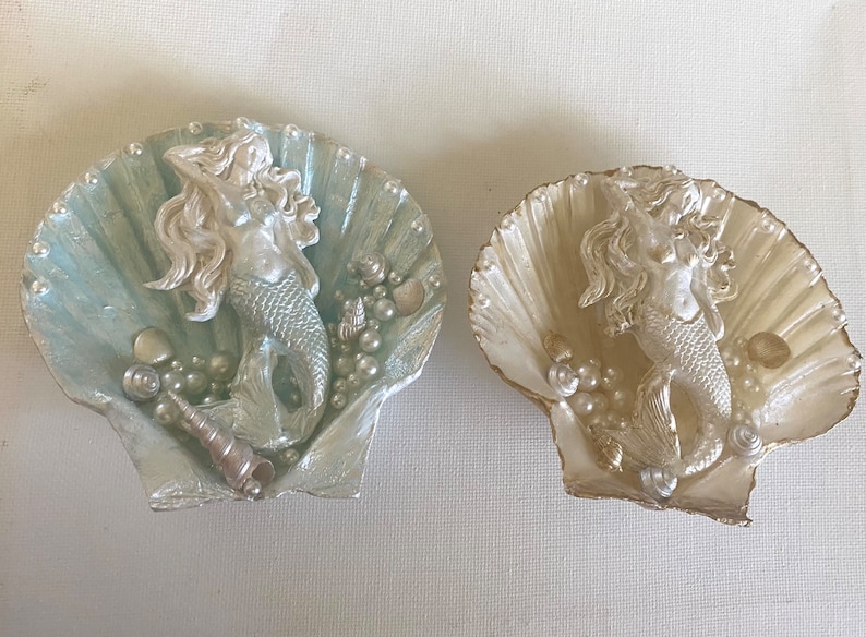 K&ouml;nnte beinhalten: Zwei dekorative Muschelschalen mit einer Meerjungfrauenfigur im Inneren. Eine Schale ist hellblau mit silbernen Akzenten und die andere ist wei&szlig; mit goldenen Akzenten.