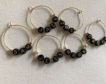 May include: Six gold-toned wine glass charms with black beads spelling out the names "Sam", "Jack", "Zoe", "Jess", "Mark", and "Cassie".