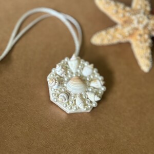 May include: A white necklace with a hexagonal pendant made of seashells and pearls. The pendant is surrounded by a circle of white pearls.