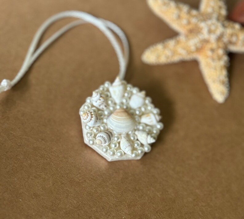 May include: A white necklace with a hexagonal pendant made of seashells and pearls. The pendant is surrounded by a circle of white pearls.