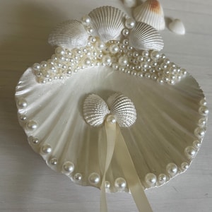 May include: A large white scallop shell decorative dish adorned with numerous small white pearls along the edges and top. Two smaller shells are attached to the center, featuring a thin cream-colored ribbon hanging down from between them.
