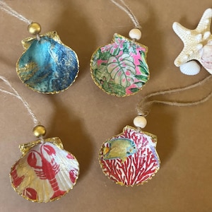 May include: Four gold-rimmed seashell ornaments with different designs. The shells are decorated with a blue and gold marbled pattern, a pink and green tropical leaf pattern, a red and white coral reef pattern, and a white and red lobster pattern.