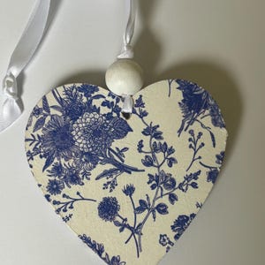 Op de afbeelding: Een hartvormige houten decoratie met een cr&egrave;mekleurige achtergrond en een blauw bloemenpatroon. Het heeft een wit lint om op te hangen en een kleine houten kraal. Een decoratief item.