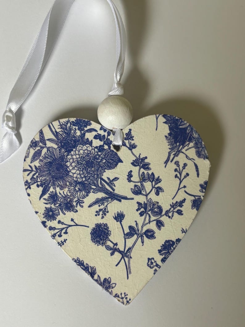 Op de afbeelding: Een hartvormige houten decoratie met een cr&egrave;mekleurige achtergrond en een blauw bloemenpatroon. Het heeft een wit lint om op te hangen en een kleine houten kraal. Een decoratief item.