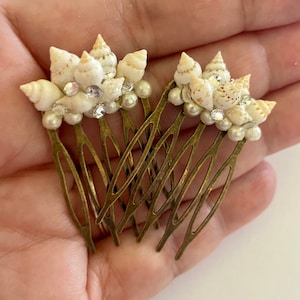 Könnte beinhalten: Zwei goldene Haarkämme, die mit weißen Muscheln und Strasssteinen verziert sind. Die Kämme werden in einer Hand gehalten.