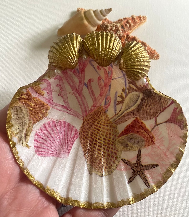 May include: A white seashell ring dish with a gold trim and a pink and brown seashell design. Three gold seashells are glued to the top of the dish, and there are two white pearl accents.