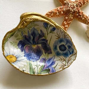 May include: A decorative seashell with a gold-painted rim. The interior features a floral design with blue irises, daisies, and other flowers. A starfish is visible in the background, suggesting a coastal theme.