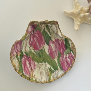 May include: A seashell with a gold trim, decorated with a floral pattern of pink and white tulips on a green background.