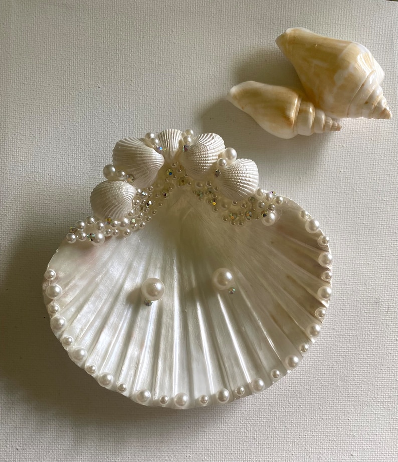 K&ouml;nnte beinhalten: Eine wei&szlig;e Muschel-Ring Schale, verziert mit Perlen und kleinen Muscheln. Die Schale steht auf einer wei&szlig;en Oberfl&auml;che.