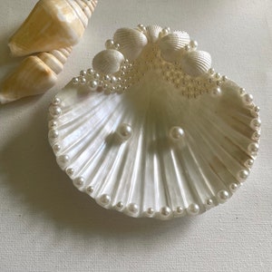 K&ouml;nnte beinhalten: Eine wei&szlig;e Muschel-Ring Schale mit einem Perlensaum und Perlenakzenten. Die Schale ist mit kleinen wei&szlig;en Perlen gef&uuml;llt und hat zwei gr&ouml;&szlig;ere Perlen in der Mitte.