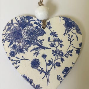 Op de afbeelding: Houten ornament in de vorm van een hart met een blauw bloemmotief op een cr&egrave;mekleurige achtergrond. Het ontwerp toont verschillende bloemen en bladeren. Een witte kraal en touw zijn bevestigd om op te hangen. Het ornament is ongeveer 10 cm breed.