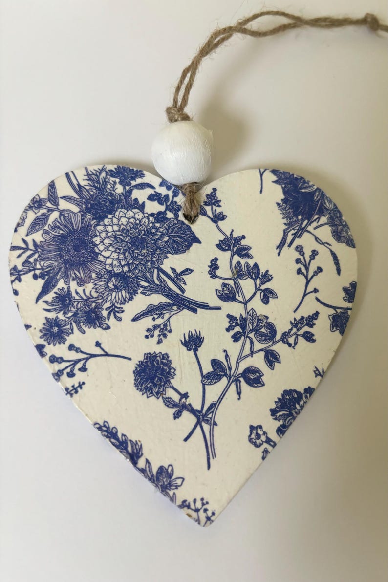 Op de afbeelding: Houten ornament in de vorm van een hart met een blauw bloemmotief op een cr&egrave;mekleurige achtergrond. Het ontwerp toont verschillende bloemen en bladeren. Een witte kraal en touw zijn bevestigd om op te hangen. Het ornament is ongeveer 10 cm breed.