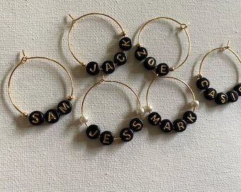 May include: Five gold hoop earrings with black letter beads spelling out names: Sam, Jacob, Zoe, Mark, and Casie. The earrings have a simple, elegant design, perfect for everyday wear.