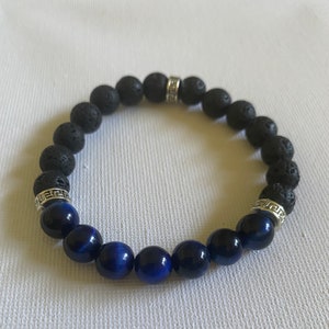 Puede incluir: Un brazalete de cuentas de lava negra y ojo de tigre azul con separadores plateados. El brazalete tiene un diseño de llave griega en los separadores.