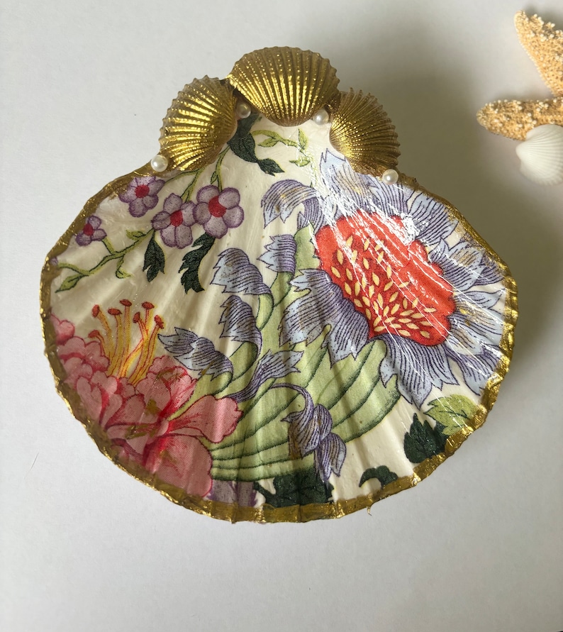 Może przedstawiać: Dekoracyjna muszelka w kształcie talerza z kwiatowym wzorem w odcieniach r&oacute;żowego, fioletowego, zielonego i ż&oacute;łtego. Muszelka jest ozdobiona złotą obw&oacute;dką i sztucznymi perełkami.