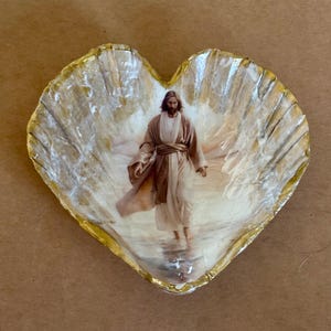May include: Heart-shaped seashell trinket dish with a gold-painted rim. The interior features a religious image of a man in a robe walking on water. The shell has a pearlescent, textured surface, creating a unique decorative piece.