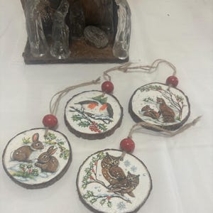 Puede incluir: Cuatro adornos redondos de rodajas de madera con un tema invernal. Cada adorno presenta un animal diferente: un petirrojo, un zorro, un par de búhos y un par de conejos. Los adornos están decorados con pintura blanca y purpurina, y cada uno tiene una cuenta roja unida a una cuerda de hilo.