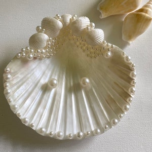 K&ouml;nnte beinhalten: Eine wei&szlig;e Muschel-Ring Schale mit einem Perlensaum und zwei Perlenohrringen im Inneren. Die Schale ist mit kleineren Muscheln und Perlen verziert.