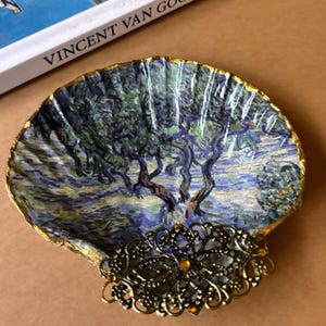 May include: A decorative seashell trinket dish featuring a painting of a tree landscape in blue, green, and yellow hues, with a gold-painted rim. A gold-colored filigree embellishment is attached to the shell. The book "VINCENT VAN GOGH" is in the background.