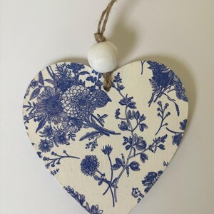 Op de afbeelding: Houten hartvormige ornament met een blauw bloemmotief op een cr&egrave;mekleurige achtergrond. Een witte houten kraal en touw zijn bevestigd om op te hangen. Het ornament heeft een klassiek, botanisch ontwerp.