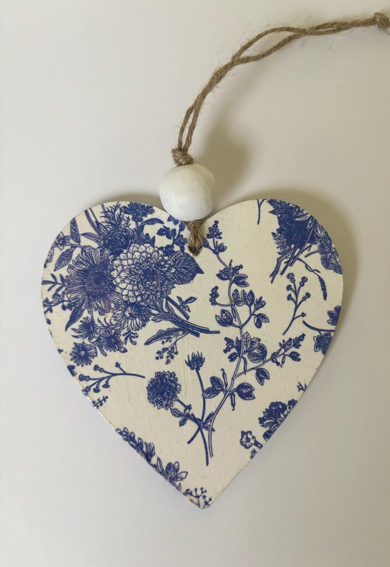 Op de afbeelding: Houten hartvormige ornament met een blauw bloemmotief op een cr&egrave;mekleurige achtergrond. Een witte houten kraal en touw zijn bevestigd om op te hangen. Het ornament heeft een klassiek, botanisch ontwerp.