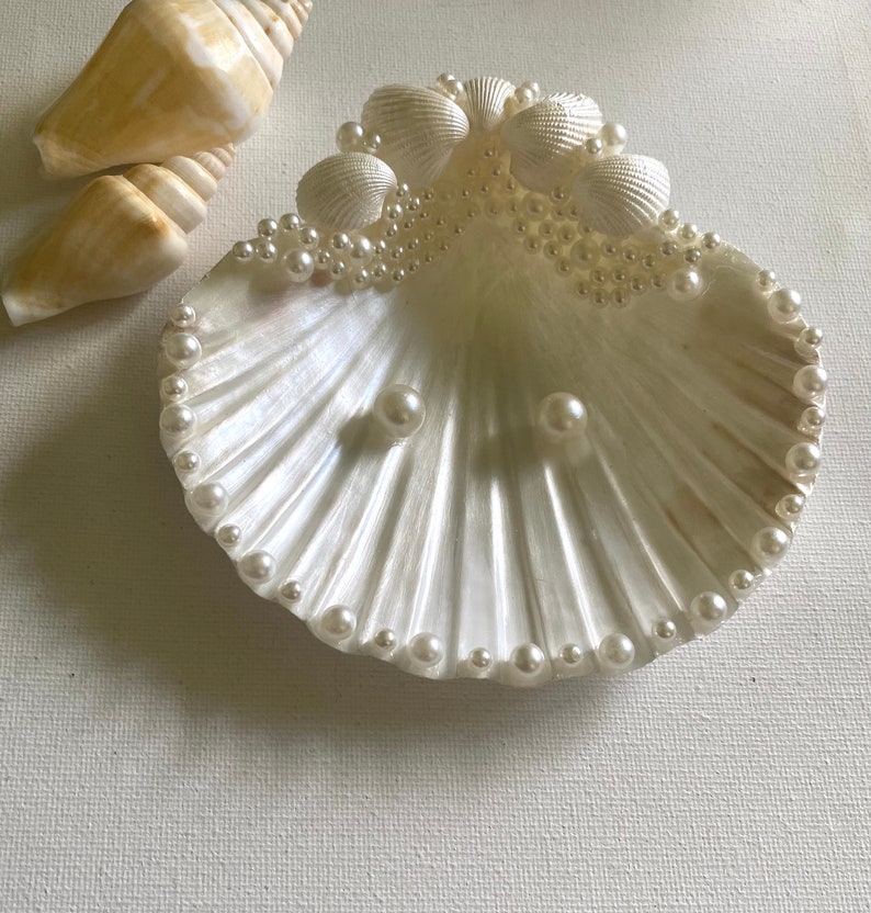 K&ouml;nnte beinhalten: Ein wei&szlig;er Muschelringhalter mit Perlensaum und Perlenakzenten. Die Muschel ist ge&ouml;ffnet und enth&auml;lt zwei Perlen.