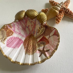 May include: A white seashell dish with a gold rim and a seashell design. The dish is decorated with pink and gold seashells, coral, and a starfish. The dish is perfect for holding jewelry or other small items.