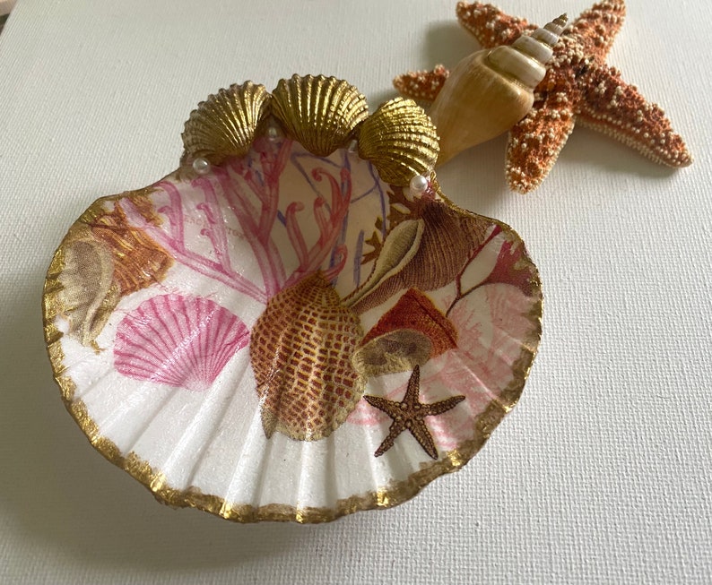 May include: A white seashell dish with a gold rim and a seashell design. The dish is decorated with pink and gold seashells, coral, and a starfish. The dish is perfect for holding jewelry or other small items.