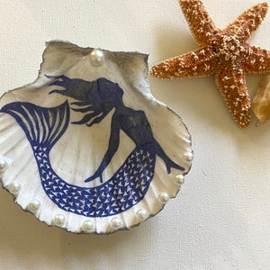 Op de afbeelding: Een witte schelp met een blauw zeemeerminontwerp en witte parel accenten. De schelp is een decoratief schaaltje.