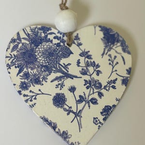 Op de afbeelding: Houten ornament in de vorm van een hart, met een cr&egrave;mekleurige achtergrond en een blauw bloemenpatroon. Bovenop is een witte kraal bevestigd met een bruin touwtje. Het ornament is een decoratief item, geschikt om op te hangen.