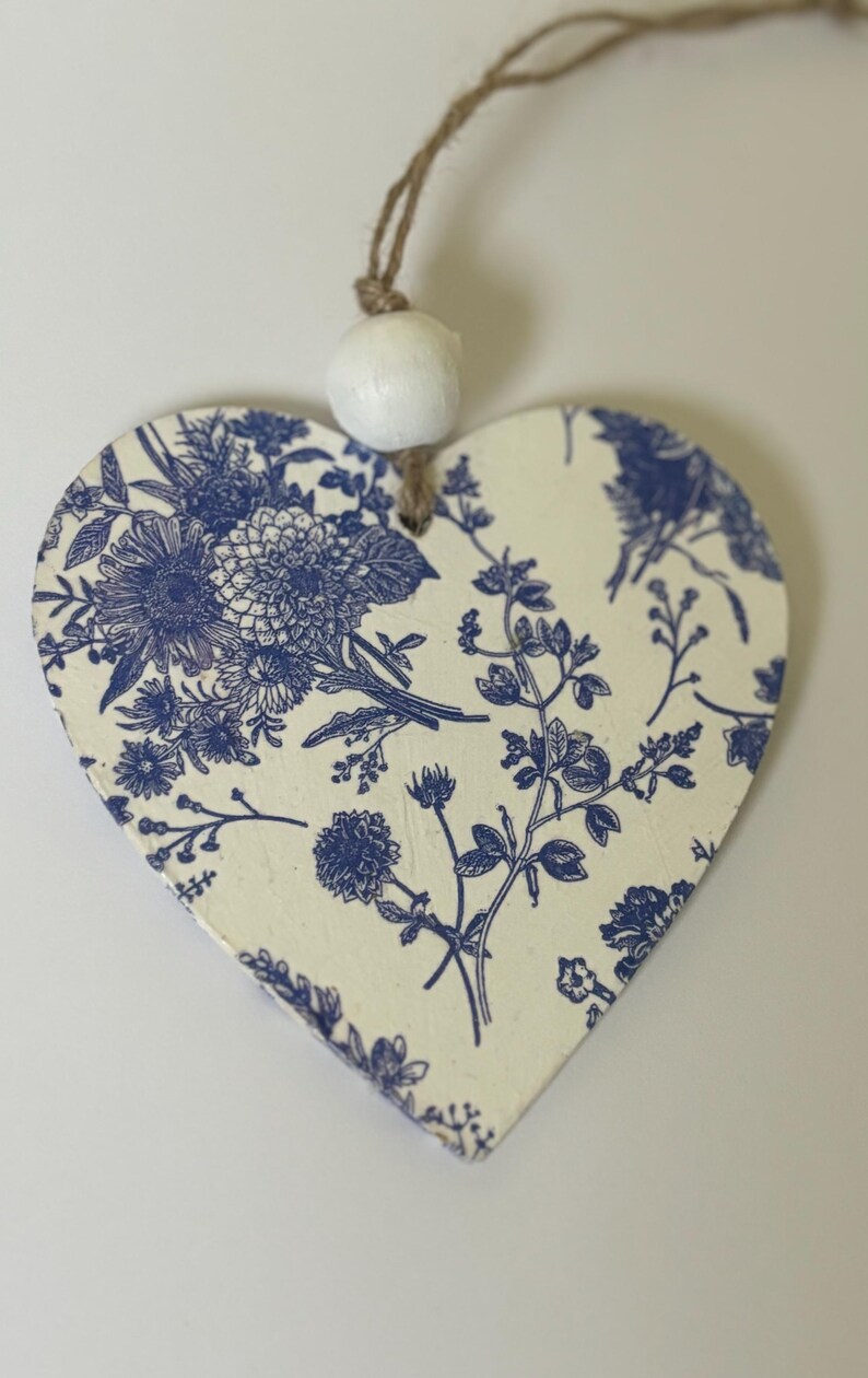 Op de afbeelding: Houten ornament in de vorm van een hart, met een cr&egrave;mekleurige achtergrond en een blauw bloemenpatroon. Bovenop is een witte kraal bevestigd met een bruin touwtje. Het ornament is een decoratief item, geschikt om op te hangen.