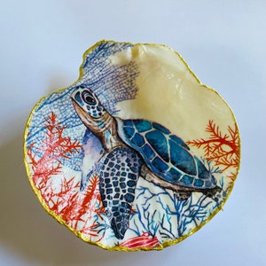 Può includere: Una conchiglia bianca con un bordo dorato, dipinta con una tartaruga marina blu e verde che nuota tra coralli e alghe.