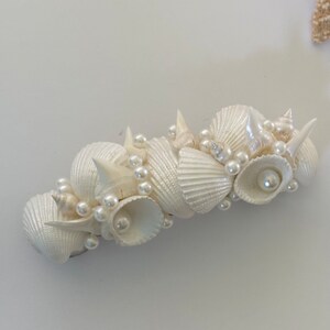 May include: A white seashell hair clip with pearl accents. The clip is made of various seashells and pearls, arranged in a cluster.