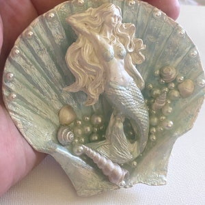 K&ouml;nnte beinhalten: Eine wei&szlig;e Meerjungfrauenfigur mit langen, flie&szlig;enden Haaren, die in einer blauen und wei&szlig;en Muschel mit Perlen- und Muschelakzenten sitzt.
