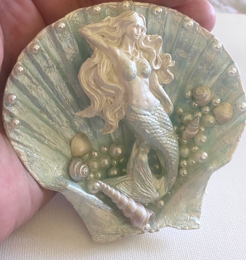 K&ouml;nnte beinhalten: Eine wei&szlig;e Meerjungfrauenfigur mit langen, flie&szlig;enden Haaren, die in einer blauen und wei&szlig;en Muschel mit Perlen- und Muschelakzenten sitzt.