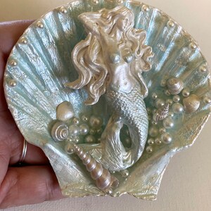 K&ouml;nnte beinhalten: Eine silberne Meerjungfrauenfigur sitzt in einer hellblauen Muschel mit Perlen- und Muschelakzenten.