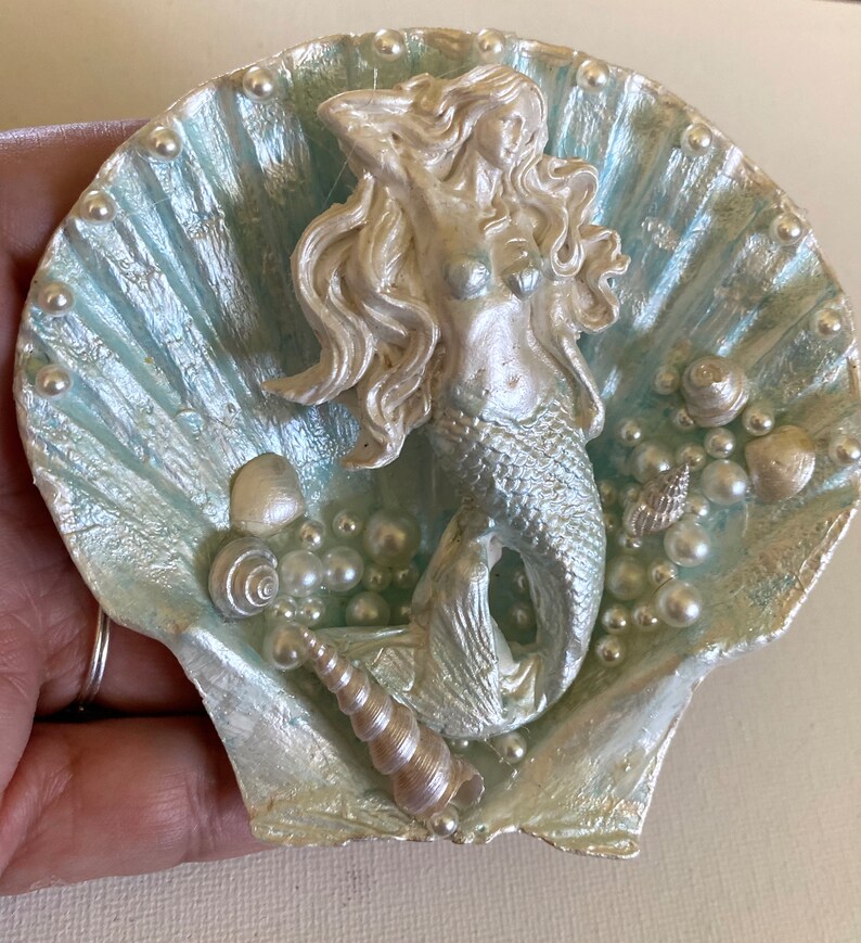 K&ouml;nnte beinhalten: Eine silberne Meerjungfrauenfigur sitzt in einer hellblauen Muschel mit Perlen- und Muschelakzenten.