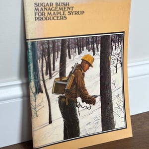 May include: A softcover booklet titled Sugar Bush Management for Maple Syrup Producers with a light orange cover. The cover features an illustration of a person in a yellow hard hat using a drill on a tree in a snowy forest setting.