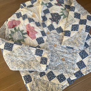 May include: A quilted jacket featuring a patchwork design of white, blue, and floral fabrics. The garment has a collared neckline, long sleeves, and a front button closure with light brown buttons. The pattern includes star shapes and squares.