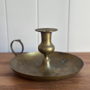May include: A brass chamberstick candle holder featuring a wide, circular drip tray base, a central stem with a bulbous middle section, a flat top candle cup, and a small, circular finger loop handle attached to the side of the tray.