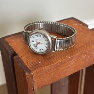 May include: A silver-toned wristwatch with a white face and black numerals sits on a wooden surface. The watch features a stretch band and a small second hand. The watch face displays the brand name "ARRAGE".