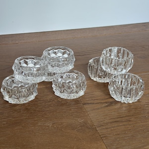 May include: A set of six clear glass napkin rings featuring a faceted geometric pattern. Each circular ring has a thick, transparent construction with a repeating diamond-shaped texture around the exterior, creating a reflective surface.