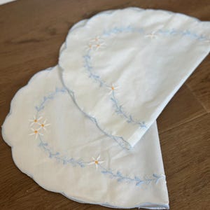 May include: Two white, scalloped-edge placemats with blue embroidered floral designs. The placemats feature a delicate pattern of small white flowers with yellow centers and blue stems. The edges are trimmed with a light blue border.